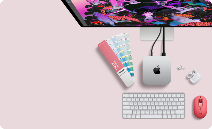 Bovenkant van Mac mini, zilverkleurig aluminium, afgeronde hoeken, glanzend zwart Apple logo in het midden, verbonden met een kabel, Studio Display deels zichtbaar aan de bovenkant, AirPods 4, opengeklapt, Pantone-kleurstalen, en Logitech-toetsenbord en -muis in rode kleur